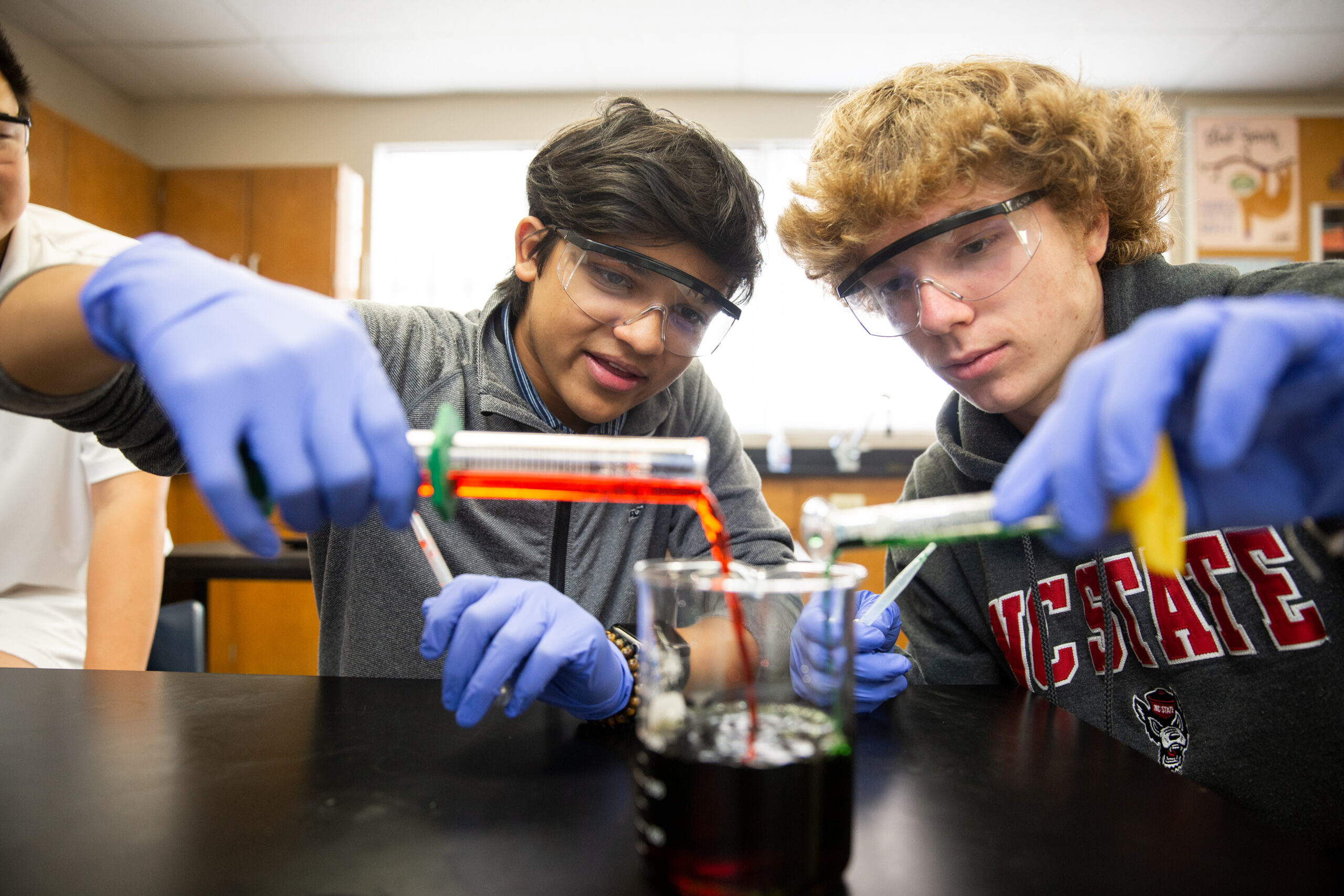 students wearing safety goggles and pouring liquid from test tubes into a beaker